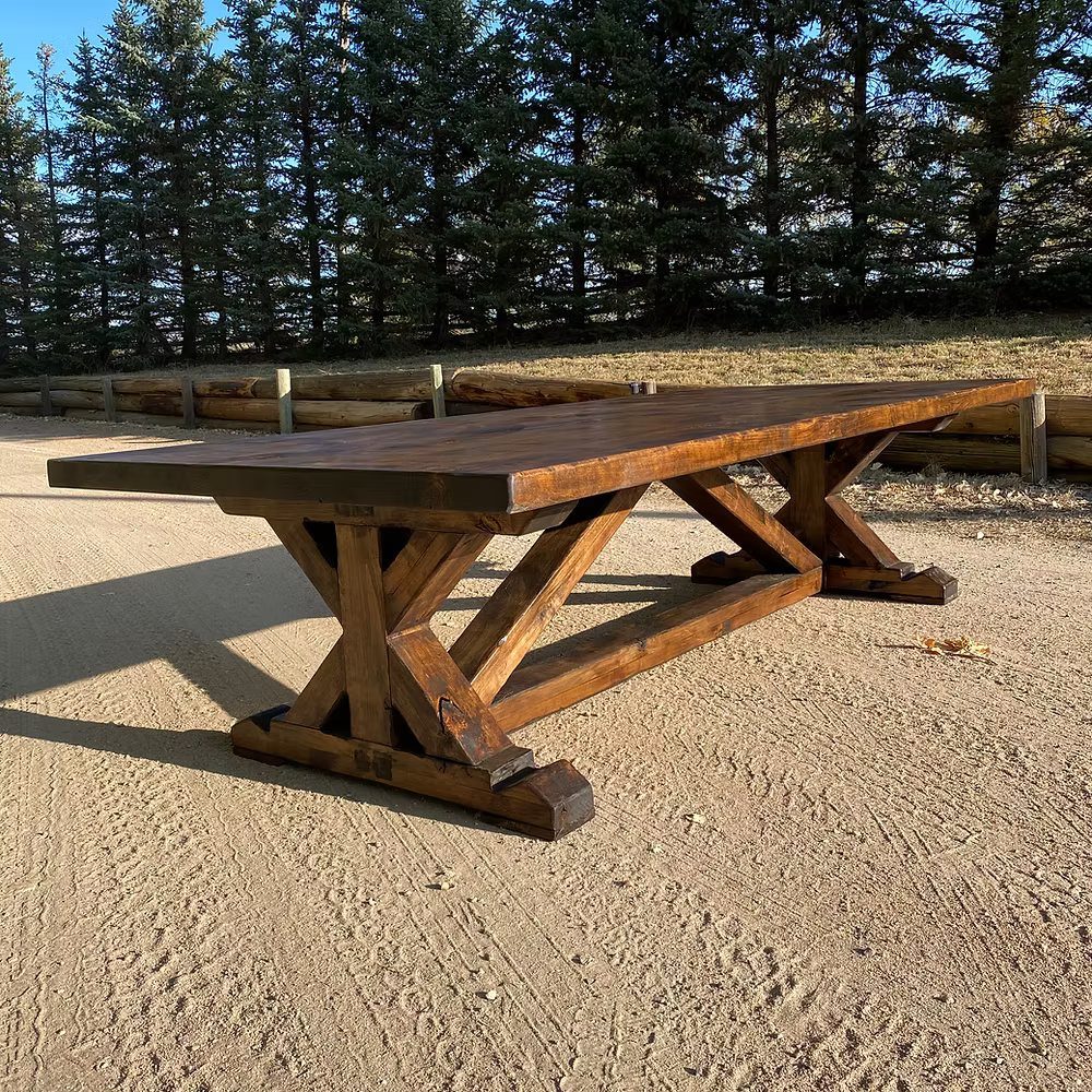 Rustic brown wooden dining table with A-frame legs standing on sandy ground outdoors.