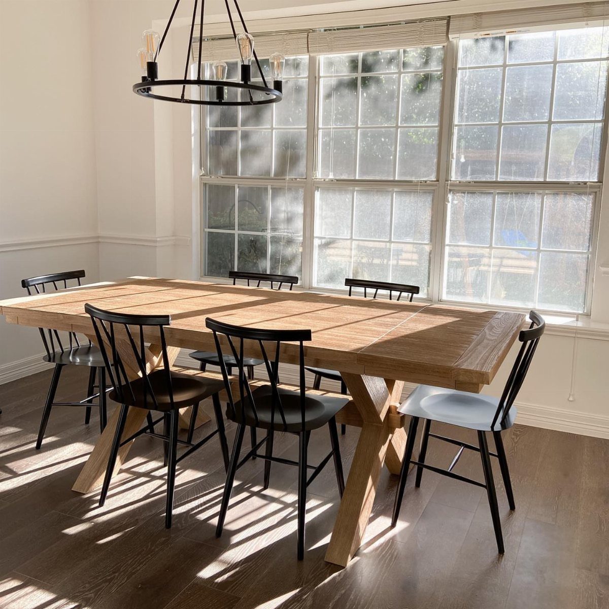 Natural wood dining table with X-legs and black metal chairs in a bright room.