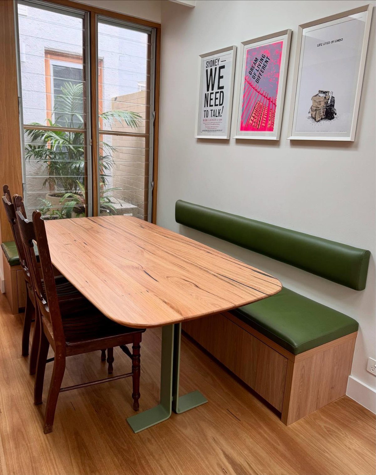 Wooden dining table with green banquette seating and dark chairs in a bright room.