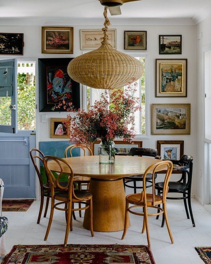 Round wooden dining table and chairs under a woven pendant lamp in an art-filled room.