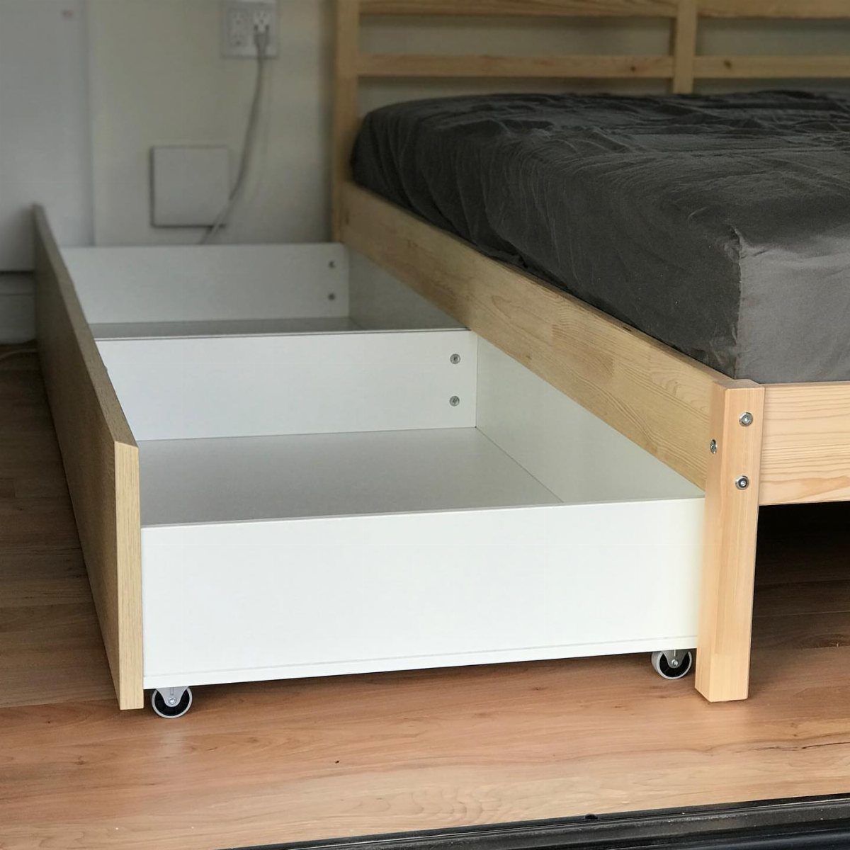 White rolling storage drawers sliding out from under a light wooden bed frame.