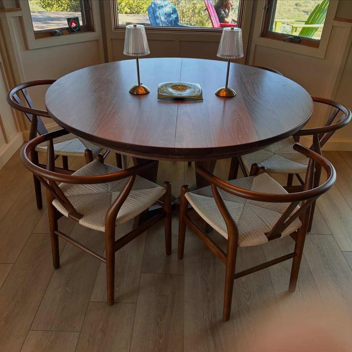 Round walnut dining table with four woven cane chairs and two small lamps.